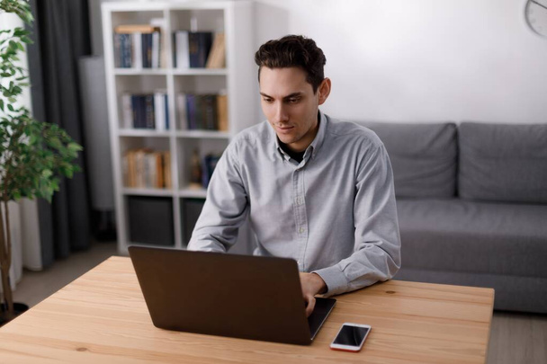 Schöner junger Mann in legerer Kleidung tippt auf einem drahtlosen Laptop, während er zu Hause im Büro sitzt. Kaukasischer Freiberufler arbeitet fernab vom gemütlichen Wohnzimmer. - Foto, Bild
