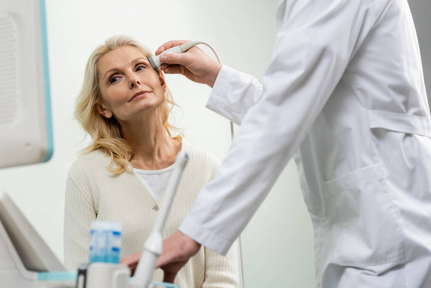 mulher loira olhando para a máquina de ultra-som enquanto médico examinando sua cabeça - Foto, Imagem
