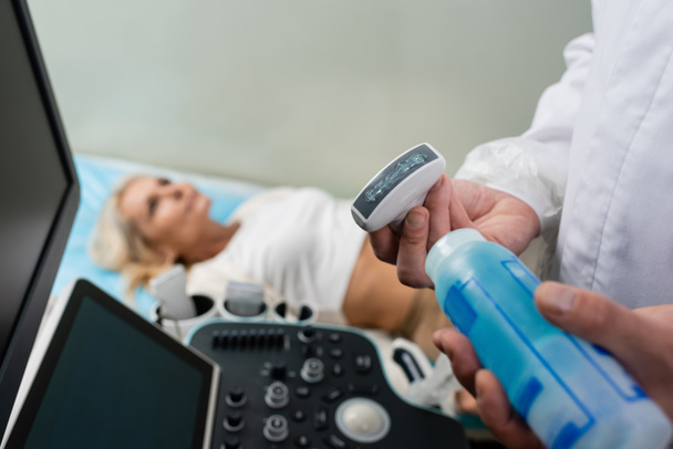 médecin appliquant un gel ultrasonique sur une sonde à ultrasons près d'un patient couché sur un fond flou  - Photo, image