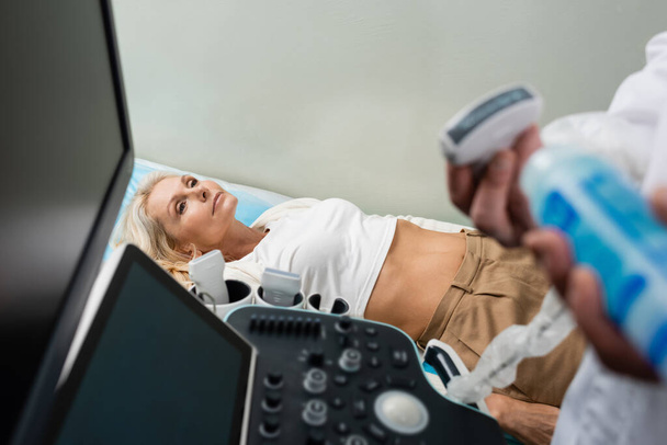 mujer de mediana edad que yace cerca de la máquina de ultrasonido y médico en primer plano borroso - Foto, Imagen