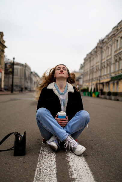 Молодая стильная женщина пьет кофе на городской улице. Европейская хипстерка со стаканом для латте на улице. Прекрасная молодая женщина с чашкой кофе на городской улице. Перерыв на кофе. Кофе в дорогу. - Фото, изображение