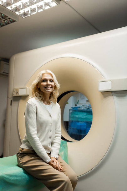 veselá zralá žena sedí v blízkosti počítačové tomografie skeneru a dívá se do kamery - Fotografie, Obrázek