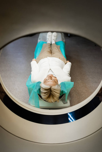 volledige weergave van middelbare leeftijd vrouw liggend tijdens onderzoek op ct scanner in kliniek - Foto, afbeelding