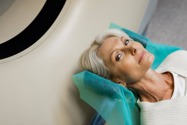 Yüksek açılı sarışın orta yaşlı kadının tomografi taraması sırasında kameraya baktığı görüntü. - Fotoğraf, Görsel