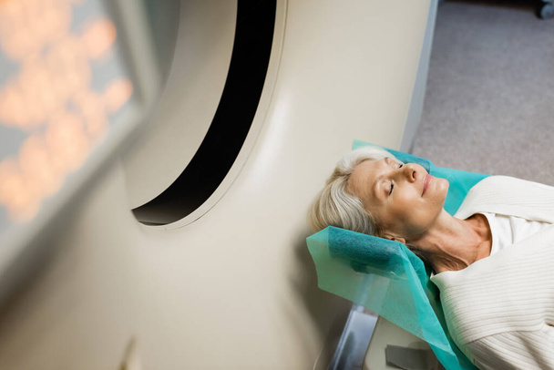 hoge hoek uitzicht van volwassen vrouw met gesloten ogen doen diagnostiek op ct scanner op wazig voorgrond - Foto, afbeelding