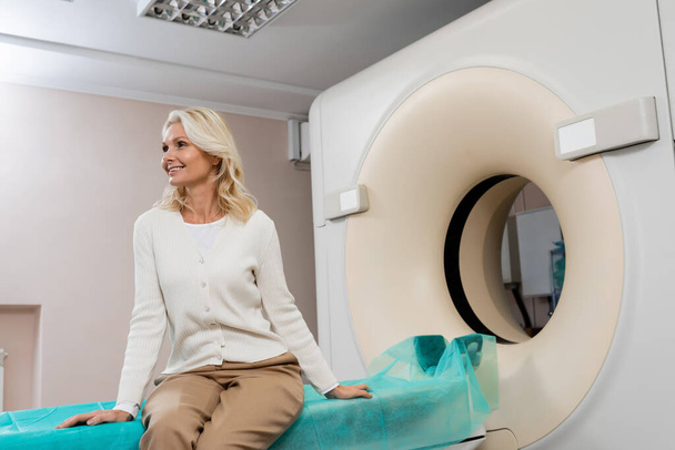 happy middle aged woman looking away while sitting near ct scanner in clinic - Photo, Image