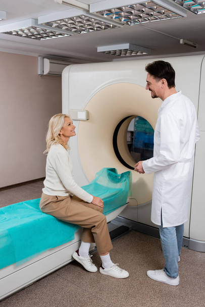radioloog in witte jas praten met glimlachende volwassen vrouw zitten in de buurt van computertomografie machine - Foto, afbeelding