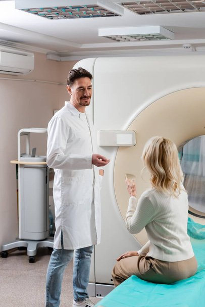 позитивный доктор в белом халате разговаривает со зрелой женщиной, сидящей рядом с компьютерным томографом - Фото, изображение