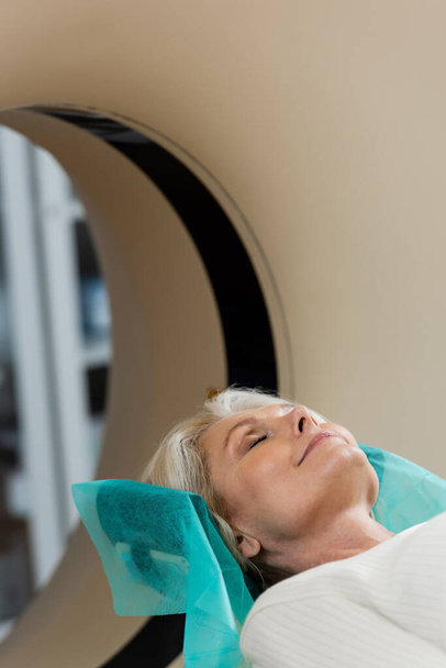 mature woman with closed eyes smiling during diagnostics on computed tomography scanner - Photo, image