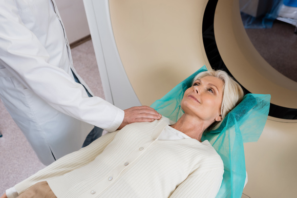 улыбающаяся женщина смотрит на рентгенолога, трогающего ее плечо перед сканированием в компьютерной томографии - Фото, изображение