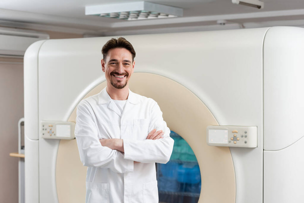 médico barbudo de bata blanca sonriendo a la cámara cerca del escáner ct en el hospital - Foto, imagen