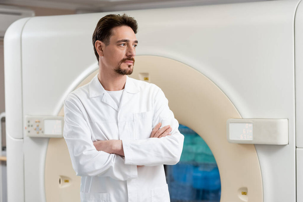 Ciddi sakallı radyoloji uzmanı klinikte tomografi cihazının yanında duruyor ve başka tarafa bakıyor. - Fotoğraf, Görsel