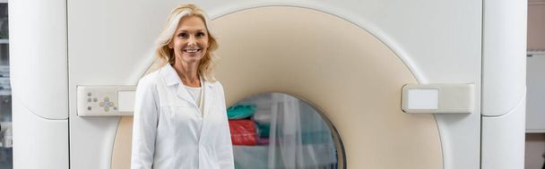 radiólogo rubia sonriente mirando la cámara cerca de la máquina de tomografía computarizada, pancarta - Foto, imagen