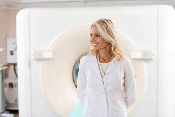 Lächelnder blonder Arzt in weißem Mantel schaut in der Nähe des Scanners im Krankenhaus weg - Foto, Bild
