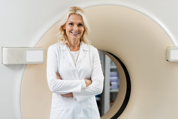 radostný lékař v bílém plášti se zkříženýma rukama poblíž počítačového tomografického skeneru - Fotografie, Obrázek