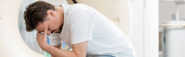 hombre frustrado cubriendo la cara mientras está sentado con la cabeza inclinada cerca del escáner de tomografía computarizada, pancarta - Foto, Imagen