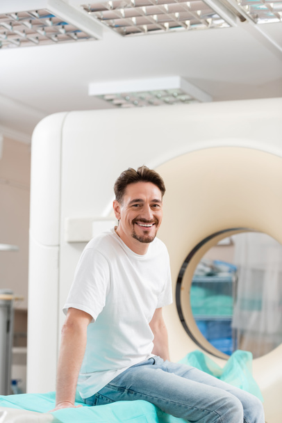 cheerful brunette man in white t-shirt sitting near ct scanner and smiling at camera - Fotografie, Obrázek