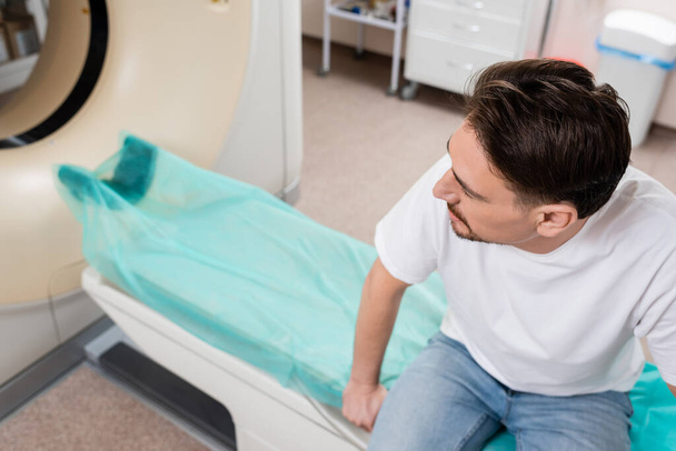 vista de ángulo alto del hombre morena en camiseta blanca sentado cerca de escáner ct en la clínica - Foto, Imagen