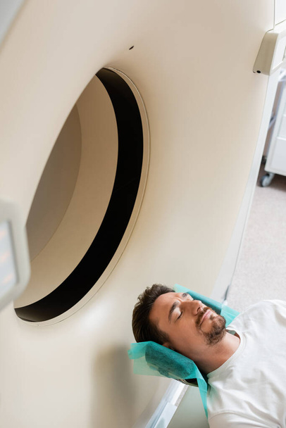 vista de ángulo alto del hombre morena acostado durante el procedimiento de escaneo en la máquina de tomografía computarizada - Foto, Imagen