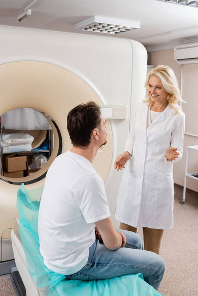radiologa bionda sorridente che parla con l'uomo bruna seduto vicino allo scanner tomografico computerizzato - Foto, immagini
