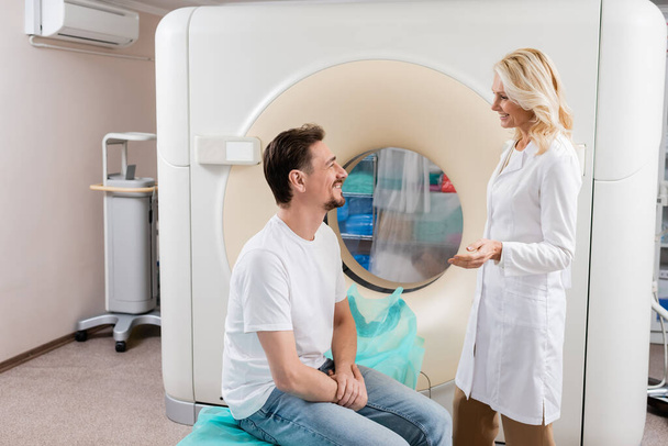 widok z boku blond radiolog rozmawia z uśmiechniętym pacjentem w pobliżu maszyny do tomografii komputerowej - Zdjęcie, obraz