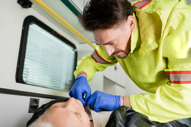 Paramédico en uniforme y guantes de látex quitándose la ropa del paciente durante los primeros auxilios  - Foto, Imagen