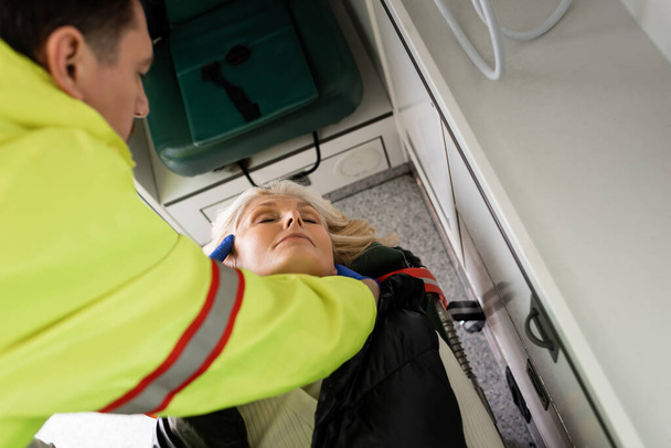 Vista de ángulo alto de paramédico borroso dando primeros auxilios a la mujer madura inconsciente en el vehículo de emergencia  - Foto, Imagen