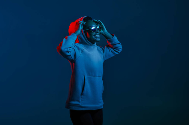 Portret zwykłej hipsterki w okularach przeciwsłonecznych i kapturze swetra na ciemnym tle pracowni w niebieskim świetle neonowym - Zdjęcie, obraz