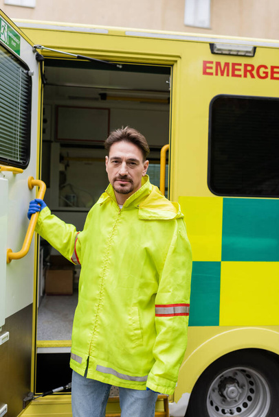 Paramédico en uniforme mirando a la cámara cerca de vehículo ambulancia en la calle urbana  - Foto, Imagen