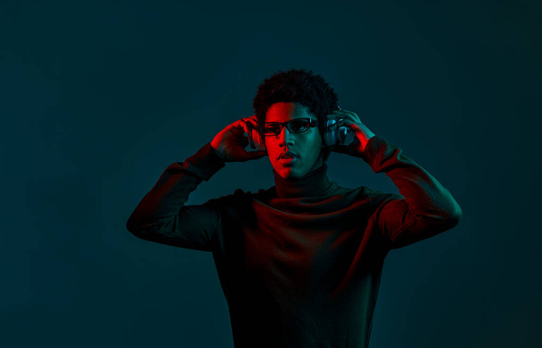 Portrait of young stylish man music lover put on headphones in neon red light over dark studio background - Fotó, kép