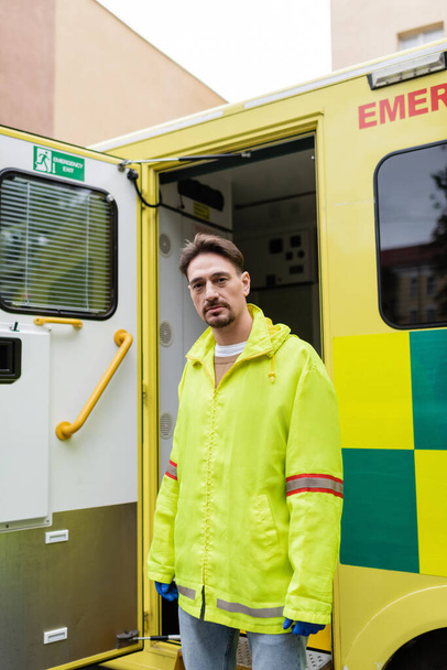 Dışarıdaki ambulans arabasının yakınındaki kameraya bakan ceketli sağlık görevlisi.  - Fotoğraf, Görsel