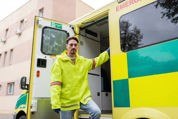 Paramédico en guantes de látex y chaqueta mirando a la cámara cerca de coche ambulancia al aire libre  - Foto, Imagen