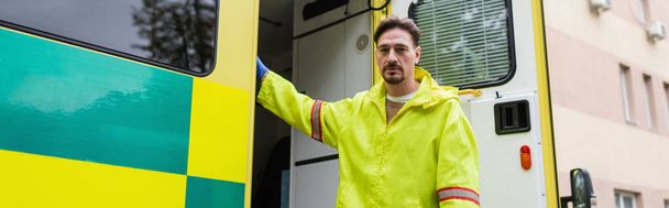 Paramedic standing near ambulance car and looking at camera outdoors, banner  - Fotoğraf, Görsel