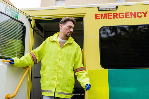 Médecin paramédical en gants de latex et veste ouvrant la porte du véhicule ambulancier à l'extérieur  - Photo, image