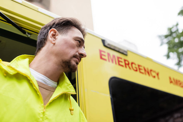 Nízký úhel pohledu na zdravotníka stojící v blízkosti rozmazané ambulance vozidla venku  - Fotografie, Obrázek
