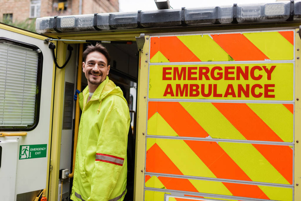 Позитивный парамедик смотрит в сторону машины скорой помощи с надписью "скорая помощь" на улице  - Фото, изображение