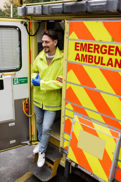 Toute la longueur du ambulancier souriant debout près du véhicule ambulancier à l'extérieur  - Photo, image