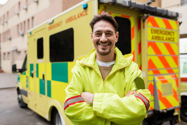 Smiling paramedic crossing arms and looking at camera near blurred ambulance vehicle outdoors  - Fotó, kép