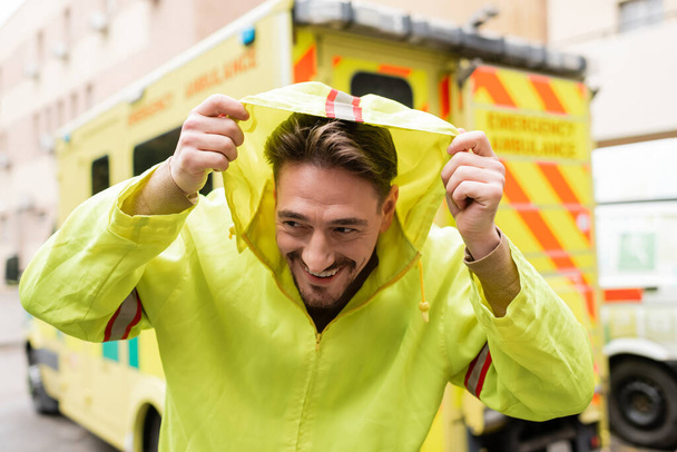 Smiling paramedic wearing hood of jacket near blurred ambulance car outdoors  - Fotografie, Obrázek