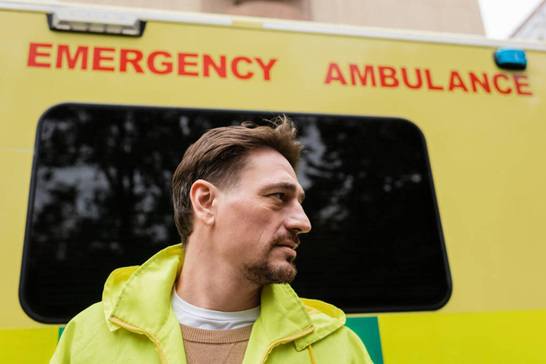 Vista lateral de paramédico mirando hacia otro lado cerca de ambulancia borrosa auto al aire libre  - Foto, Imagen