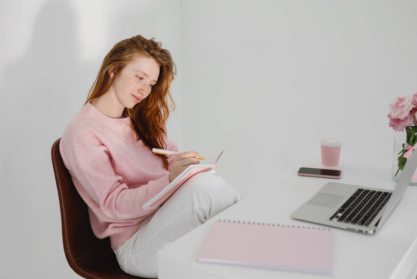 Relaxed pensive young redhead woman sitting at workplace in office and making notes. - Φωτογραφία, εικόνα