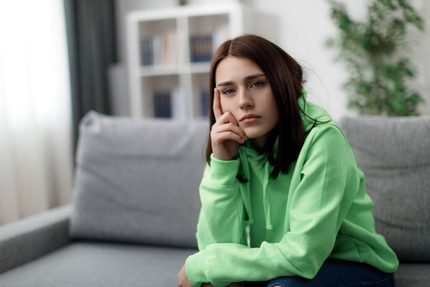 Kavkazská mladá žena se smutným výrazem ve tváři, sedící sama na gauči. Nenuceně oblečená brunetka ztracená v myšlenkách během volného času doma. - Fotografie, Obrázek