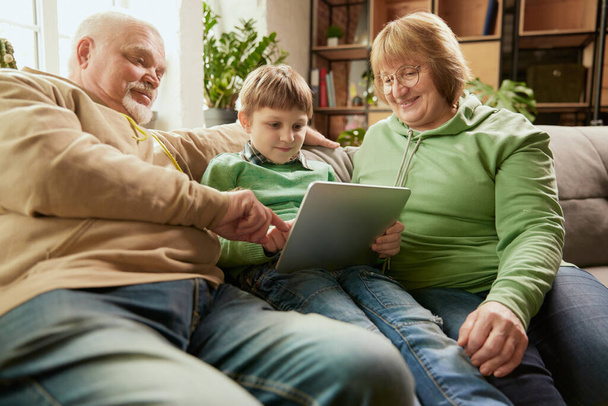 Happy friendly family, grandparent and grandson sitting on sofa and spending time together, using modern gadgets, talking, studying. Concept of love, care, support, hobbies and leisure activities - 写真・画像