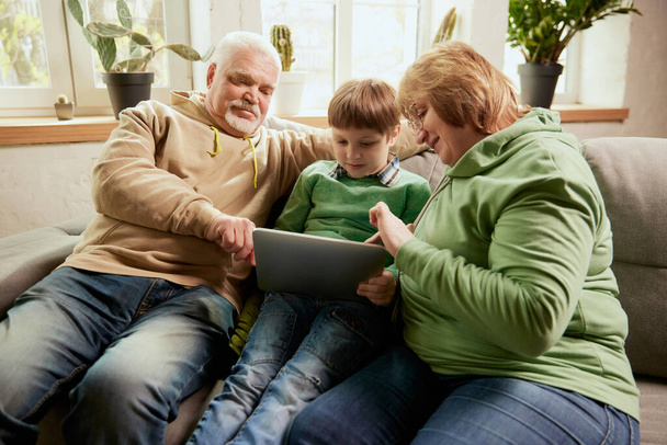 Life portrait of friendly family, grandparent and their grandson sitting on sofa and spending time together, using modern gadgets, talking, studying. Concept of love, care, support, hobbies and - Фото, зображення