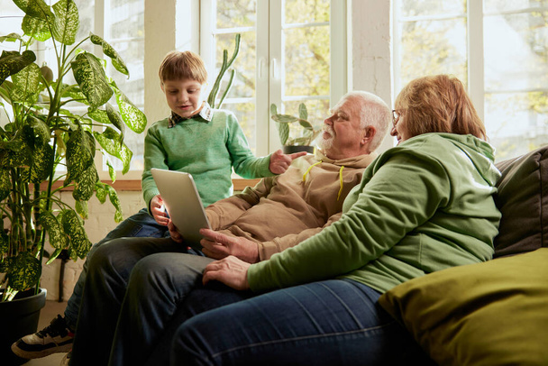 Happy friendly family, grandparent and grandson sitting on sofa and spending time together, using modern gadgets, talking, studying. Concept of love, care, support, hobbies and leisure activities - Zdjęcie, obraz