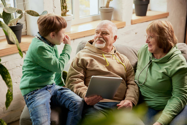 Old grandparents with grandchild sitting on couch at home and reading news, funny stories. Watching reels at laptop. Happy time in friendly and cozy atmosphere. Concept of family, love and education - Foto, Imagem