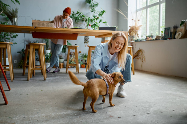 Young people working at cozy florists workshop. Focus on smiling kind woman caressing cute dog while hipster man creating plant composition standing at table - Фото, изображение