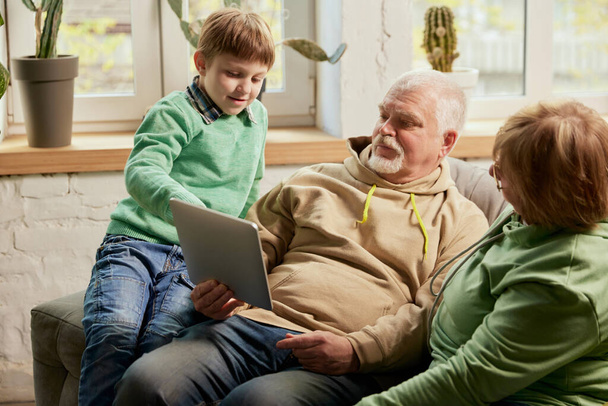 Life portrait of friendly family, grandparent and their grandson sitting on sofa and spending time together, using modern gadgets, talking, studying. Concept of love, care, support, hobbies and - Fotoğraf, Görsel