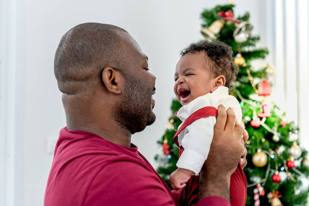 Африканская семья, 4-месячный нигерийский младенец новорожденный сын и отец, улыбающиеся и счастливые вместе, с размытым фоном рождественской елки, африканской семье на праздник и ребенка концепции младенца. - Фото, изображение