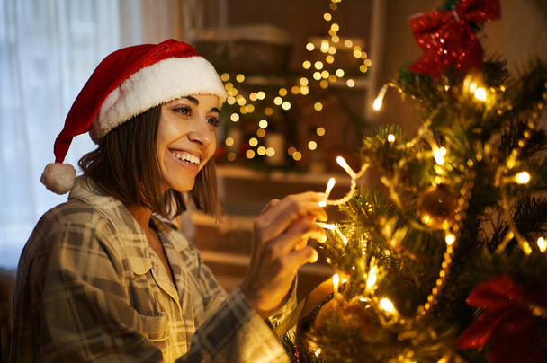 Joyful Santa Woman preparing for the Christmas holiday, decorating a Christmas tree with lights and toys - Photo, Image